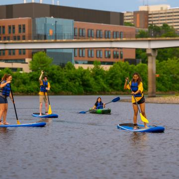 Students paddle board on the Chippewa River with the newly-constructed Science and Health Sciences building in the background.