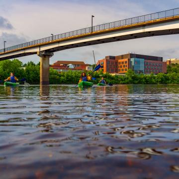 Students kayak on the Chippewa River with the Science and Health Sciences building and the bridge in the background.