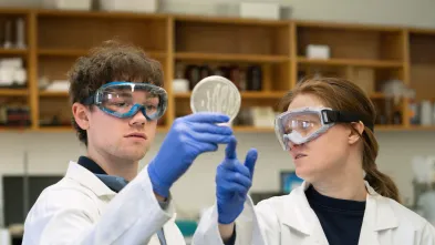 Two students hold up a petri dish and discuss what they see. The students are wearing safety glasses and coats.