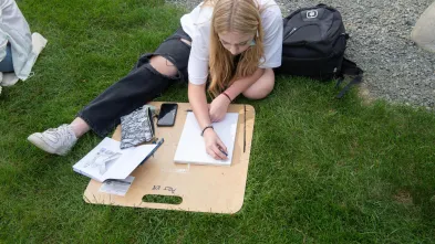 Student draws using a clipboard in the grass for an outdoor art class