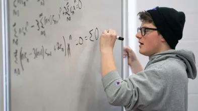 Student in Blugold sweatshirt solves a math problem on a whiteboard.