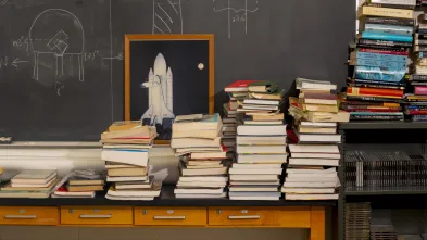 A stack of physics textbooks in front of a chalkboard and an image of a rocketship.