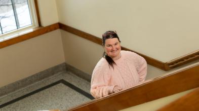 Woman with brown hair and pink sweater standing in staircase in Schofield Hall