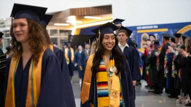 Students wearing graduation caps and gowns smile as they walk in to the ceremony