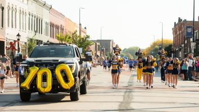 A fun view of the Homecoming parade featuring a car with the number "100" in balloons on the front while people march alongside.