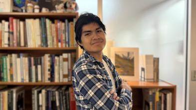 Honors student Kevin Miro stands smiling with arms crossed in front of bookshelves