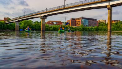 Students kayak on the Chippewa River with the Science and Health Sciences building and the bridge in the background.