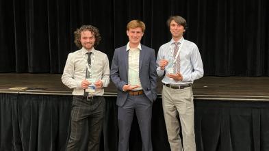 3 male students standing holding awards near a stage