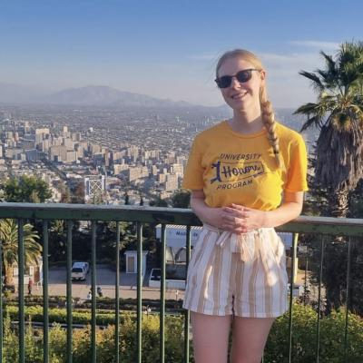 A young woman in an Honors t-shirt stands in front of a city scape.