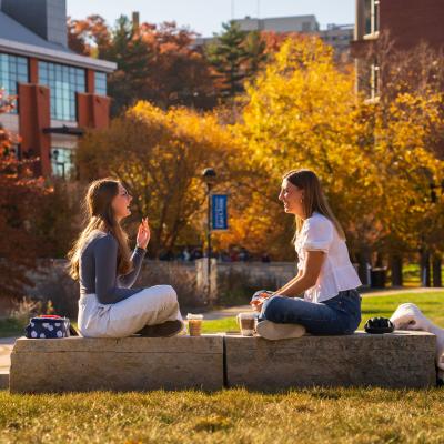 Two students sit on a rock on campus mall, laughing, talking, and drinking coffee