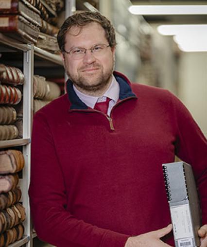 Profile images of Greg Kocken, University Archivist, standing inside inside the archival storage area while holding a box under one arm.