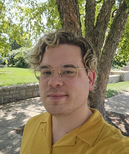 An image of Professor Chris Outzen, a white cisgender man in his mid-30s with blonde hair, in a yellow shirt under a tree. 