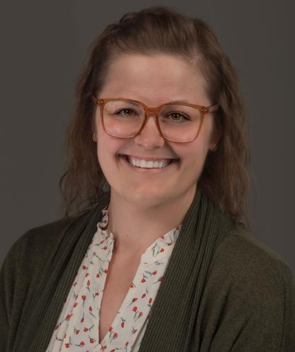 Person with glasses wearing a green cardigan and floral blouse against a gray background.