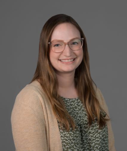 White woman with brown hair and clear glasses wearing a green shirt and cream cardigan.
