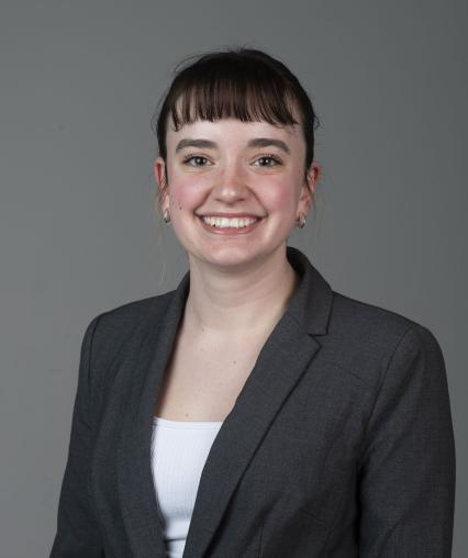 Image of a grey background, young white woman with brown hair, a white shirt, and grey blazer smiling at the camera.