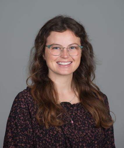 A smiling young woman with long, wavy brown hair and green-framed glasses, wearing a black floral-patterned top against a gray background.