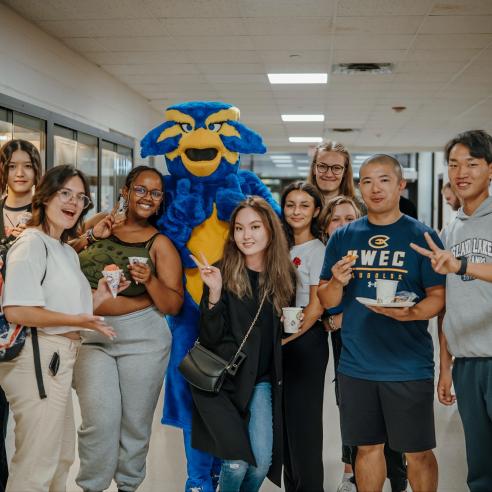 A group of students posing with a blu mascot in a library hallway, smiling and holding snacks, during a Library after dark event