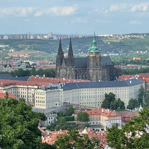 An aerial view of Prague showing the historic Prague Castle with its Gothic spires, surrounded by modern cityscape and lush greenery.