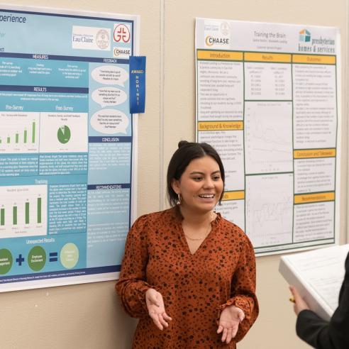 A student presenting her research at a CHAASE event, engaging in a discussion with another attendee in front of various displayed research posters.