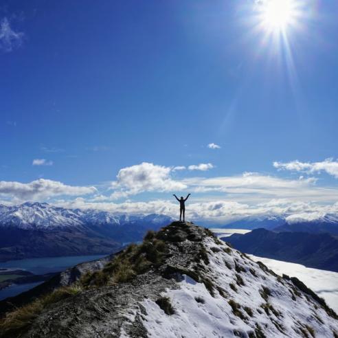 A student on a study abroad program in New Zealand standing at the peak of mountain with a large landscape mountain background