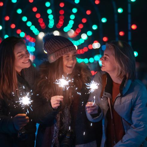 Three women holding sparklers, laughing and enjoying themselves at a nighttime outdoor event, with colorful bokeh lights in the background.