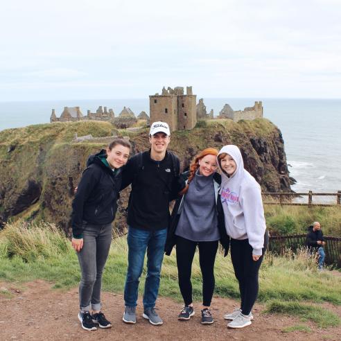 A group of students study aboard in Scotland posing together in front of the historic ruins of Dunnottar Castle
