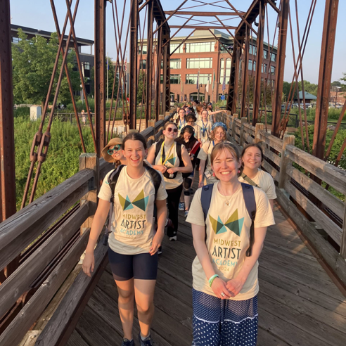 Students wearing "Midwest Artist Academy" shirts smile as they walk across a bridge