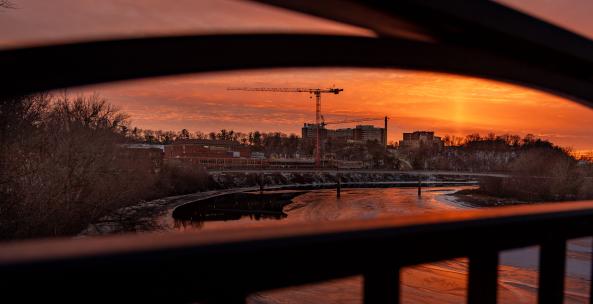View of a bright orange sunset and campus buildings in between railings of the bridge. 
