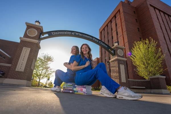 two nursing grads in scrubs sitting on textbooks under the archway 