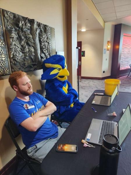 UWEC student Jack Petrizzo sitting arms crossed at a folding table next to Blu the Blugold mascot