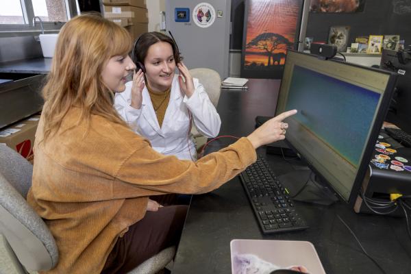 Two female students sitting side by side, in lab coats, looking at a monitor screen, one is pointing at a spot on the screen