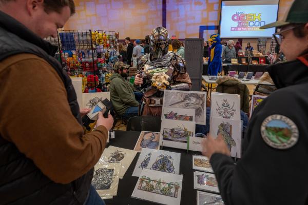 Costumes and art on display GEEKcon visitors look at art for sale. A Mandalorian cosplay is visible in the background.