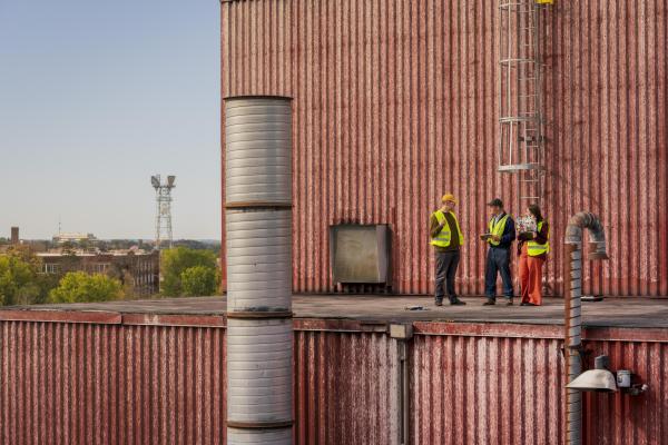 three people on top of industrial building in safety gear, yellow vests and hard hats