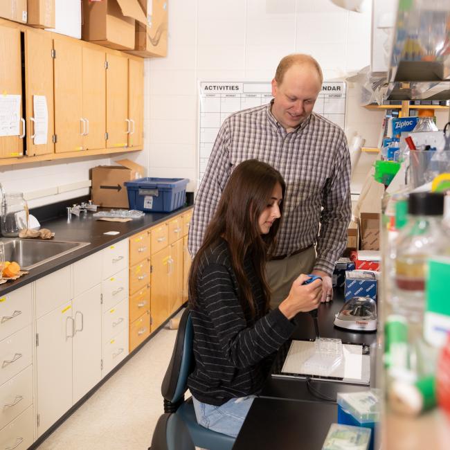 faculty person standing next to a seated student working in a chemistry lab 