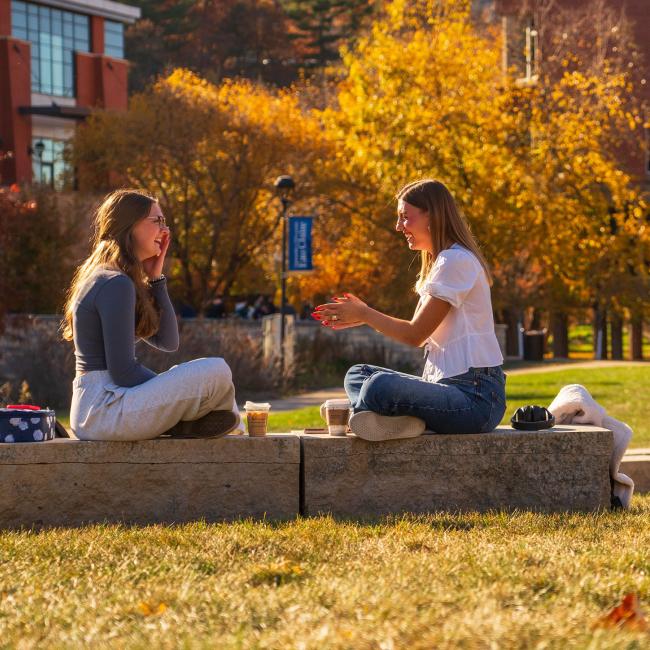 two students outside on the mall in the sunshine, fall, seated on bench