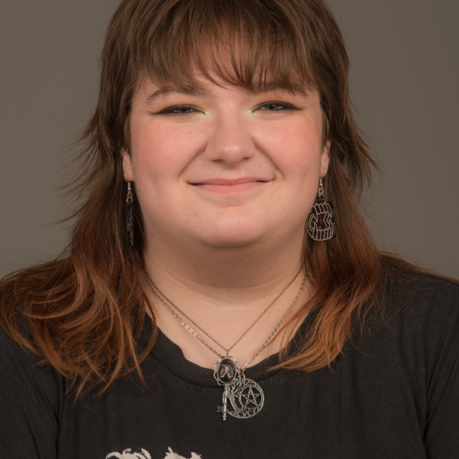 A nonbinary person with brown shoulder length hair and bangs. They are wearing an assortment of silver jewelry and a band T-shirt. They are smiling.