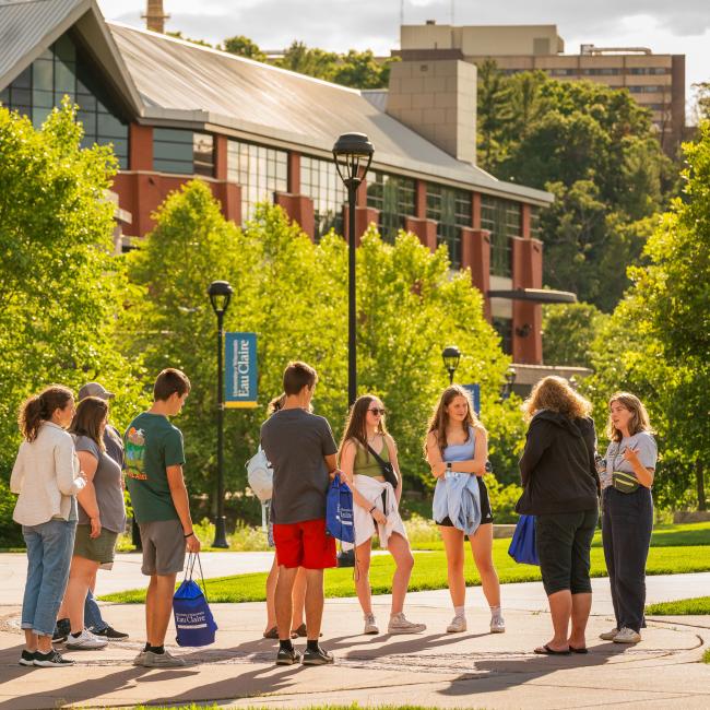 Students and parents gather on a campus sidewalk during a tour on a sunny day
