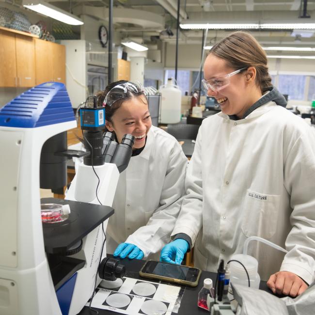 Two students wearing white coats laugh during a Biomedical Engineering Lab surrounded by equipment.