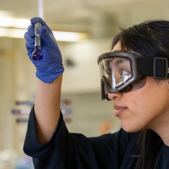 A student wearing safety goggles closely examines liquid in a small test tube