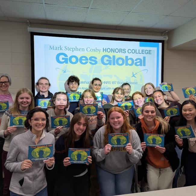A group of students hold booklets in front of a computer display.