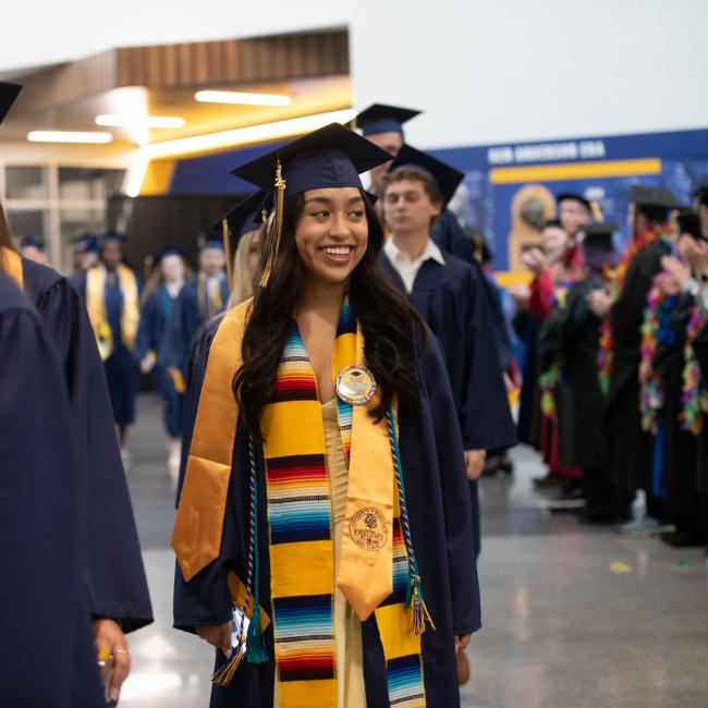 Students wearing graduation caps and gowns smile as they walk in to the ceremony