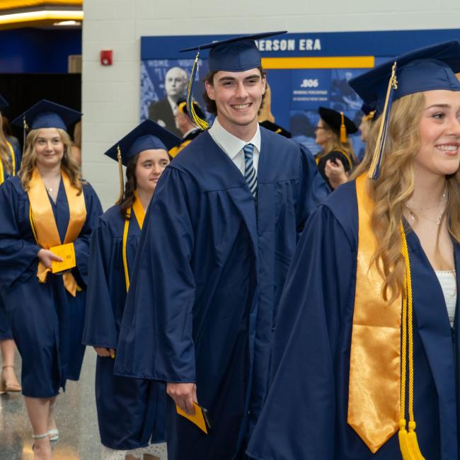 Graduates wearing graduation caps and gowns smile as they walk into the ceremony on commencement day