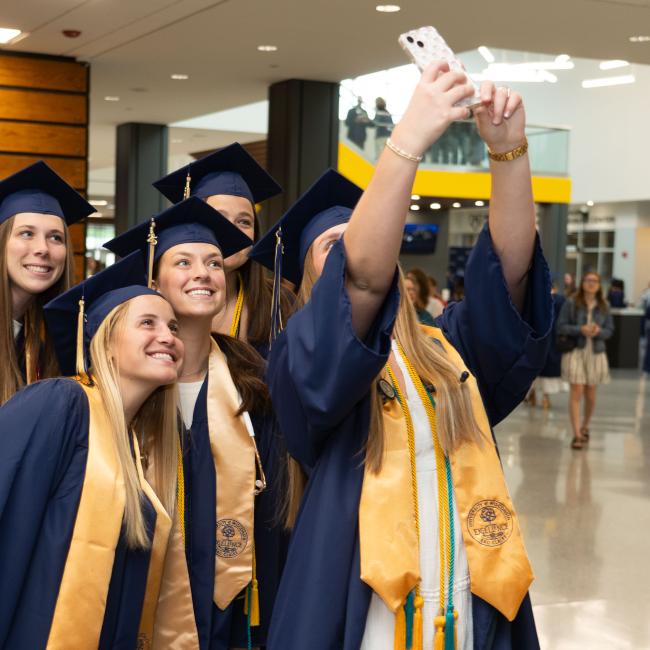 A group of grads pose for a selfie wearing graduation caps and gowns