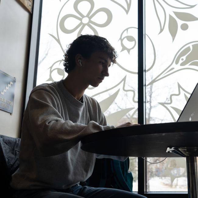 A student studies on his laptop at a table by a window.