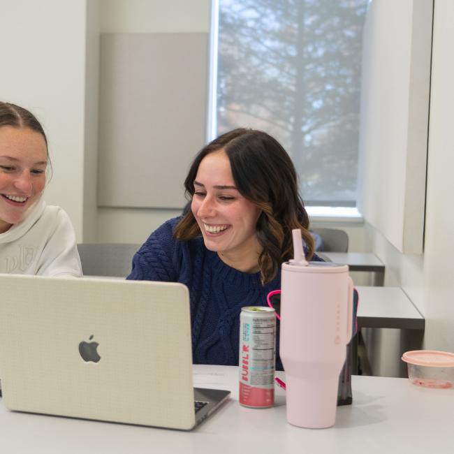 Two students smile and point to their computer while working in a classroom.