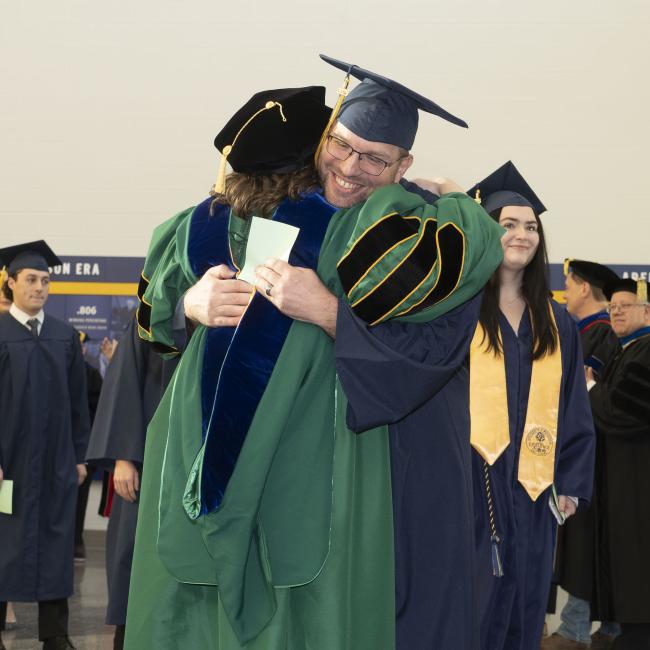 A student smiles while hugging a professor during Commencement.