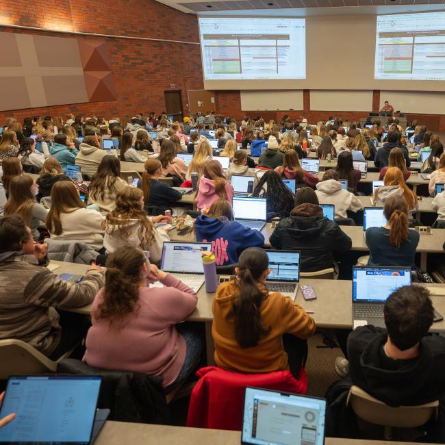 A large lecture hall features seats filled with students learning from a professor.