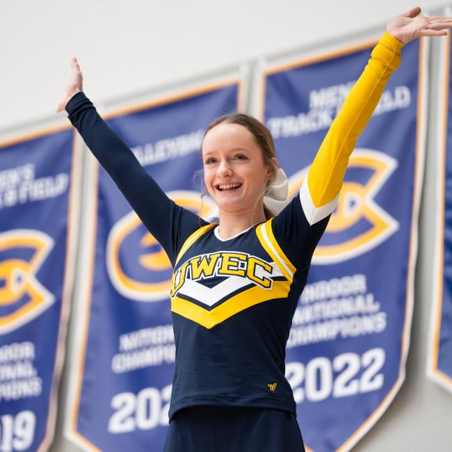A cheerleader puts her hands in the air against a backdrop of UWEC banners.