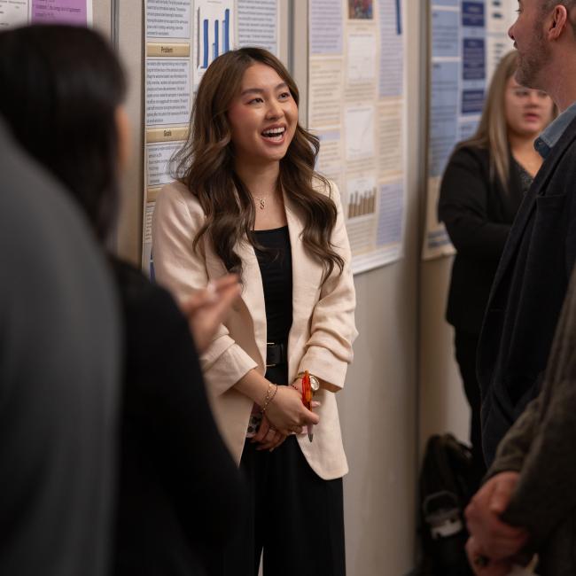 An HCAD student smiles while standing in front of their project poster and speaking with event goers