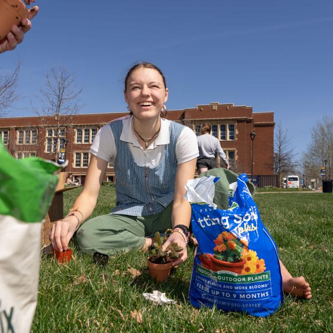 A student smiles while sitting in the grass and potting a plant on Earth Day